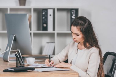 İşyerinde oturan ve not defterinde yazılı konsantre genç iş kadını