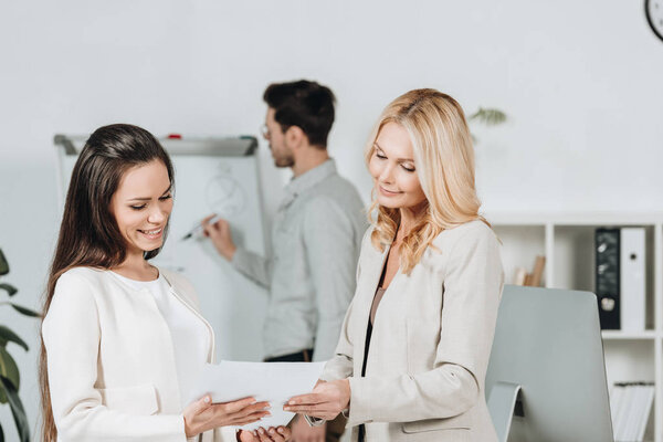 beautiful smiling businesswomen holding papers and young businessman writing on whiteboard behind in office