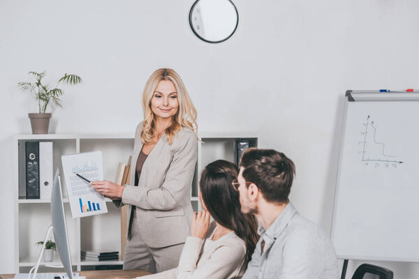 smiling business mentor pointing at charts and looking at young colleagues sitting in office
