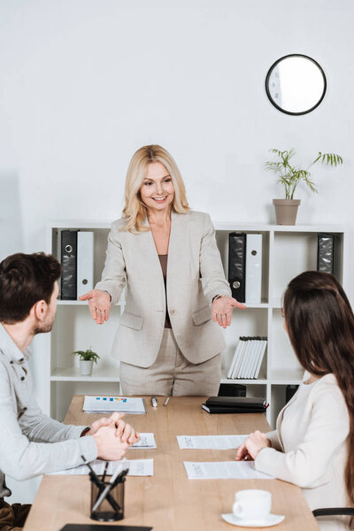 smiling female business mentor looking at young colleagues sitting at workplace