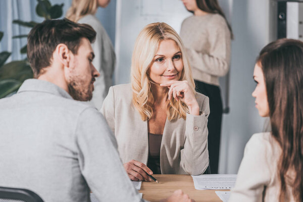 beautiful smiling business mentor sitting with hand on chin and looking at young colleagues in office
