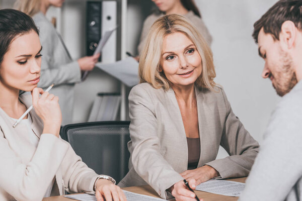 улыбающаяся зрелая деловая женщина, указывающая на документы и работающая с молодыми коллегами в офисе
