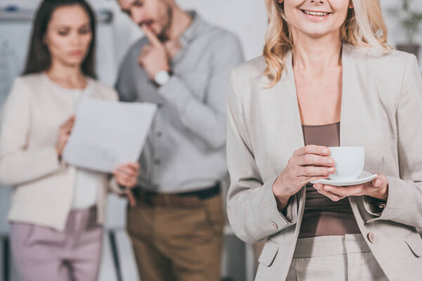 cropped shot of smiling businesswoman holding cup of coffee and young colleagues working behind in office