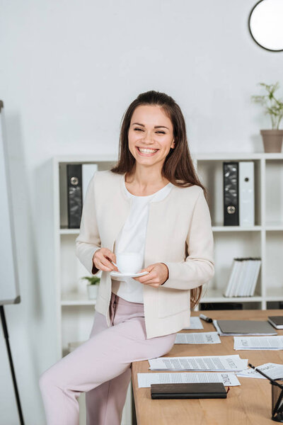 beautiful happy young businesswoman holding cup of coffee and smiling at camera in office