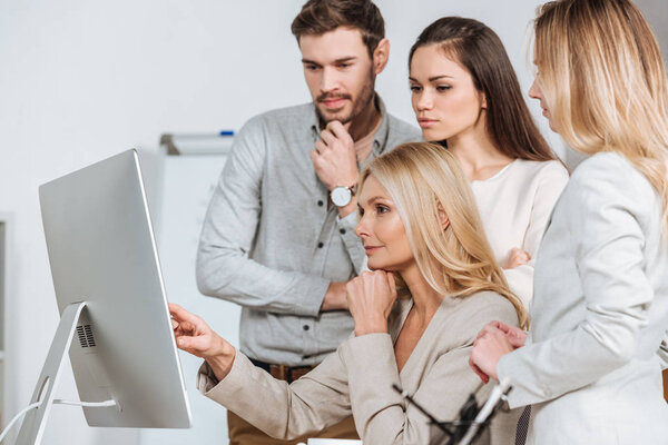 mature business mentor pointing at desktop computer and working with young colleagues in office