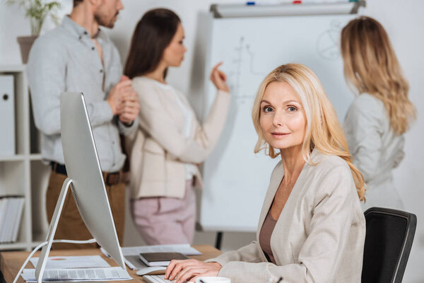 mature businesswoman using desktop computer and looking at camera while young colleagues working with whiteboard behind