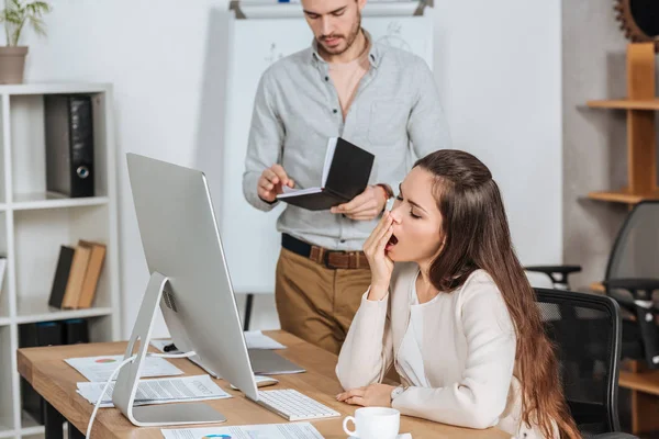 business mentor holding notebook and young female colleague yawning at workplace