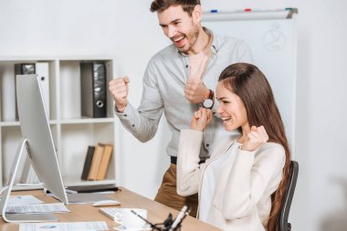 happy young business colleagues shaking fists and looking at desktop computer in office clipart