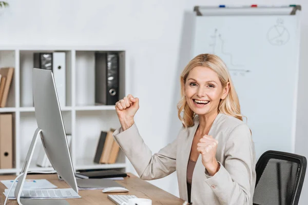 excited businesswoman shaking fists and smiling at camera while sitting at workplace