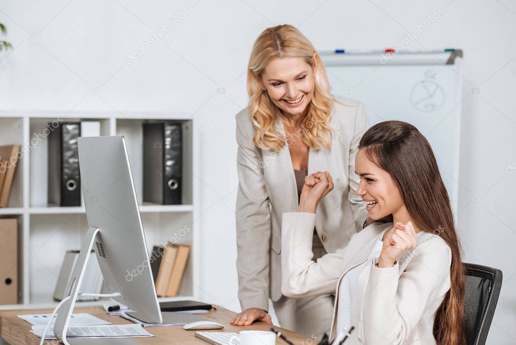 smiling mature businesswoman looking at cheerful young colleague using desktop computer at workplace