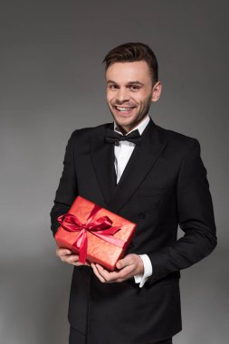 Genç adam siyah smokin ve gri izole kırmızı hediye kutusu tutarak kravat yay gülümseyen