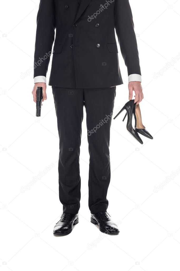 cropped view of secret agent in black suit holding handgun and high heels, isolated on white