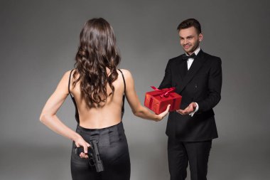 woman gifting red present to man while hiding gun behind the back, isolated on grey