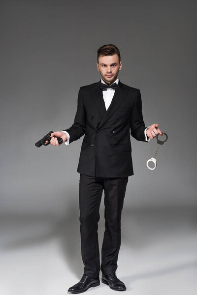 elegant secret agent in tuxedo holding handgun and handcuffs on grey