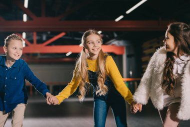 Selective focus of smiling beautiful children holding hands while moving on roller rink clipart
