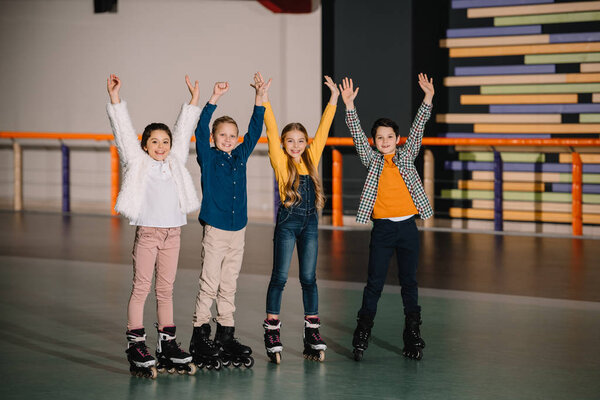 Happy smiling children in roller skates standing in spacious roller rink with hands up