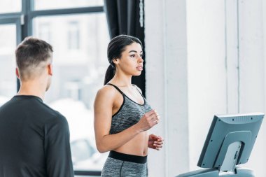 young man looking at sporty african american girl running on treadmill in gym