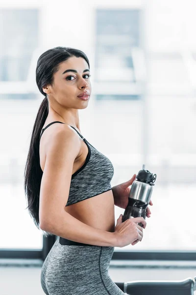 美丽的非洲裔美国女运动员拿着瓶子 看着相机在健身房 — 图库照片
