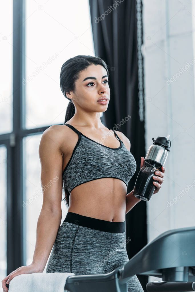 pensive young african american woman standing on treadmill and holding sport bottle