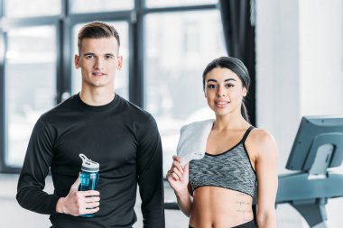su şişesi ve havlu ile güzel afro-amerikan kadın ile yakışıklı adam birlikte ayakta ve spor salonunda kamera gülümseyerek