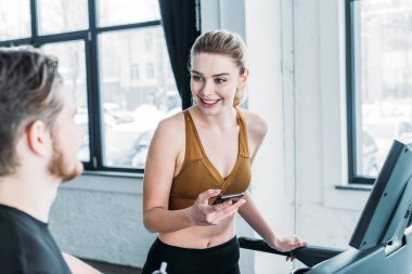 smiling girl exercising on treadmill with smartphone in hand and looking at young man on foreground in gym