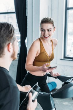 smiling sporty girl running on treadmill and looking at young man on foreground in gym