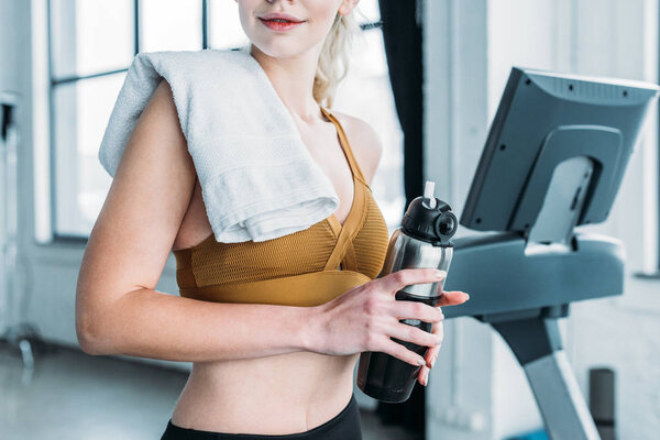 cropped shot of young sportswoman with towel on shoulder holding sports bottle in gym