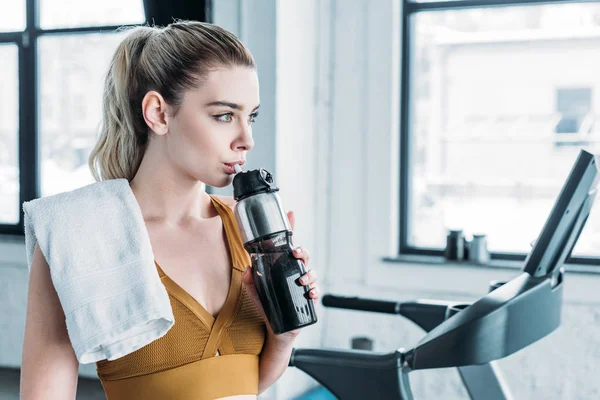 美丽的女运动员与毛巾在肩膀上饮用水从运动瓶在健身房 — 图库照片