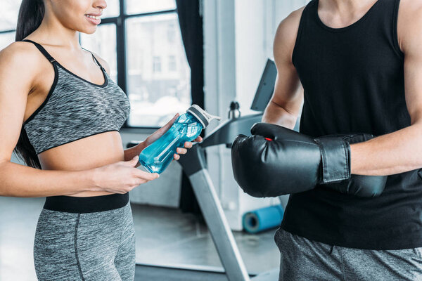 cropped shot of young man wearing boxing gloves and african american woman holding sports bottle in gym