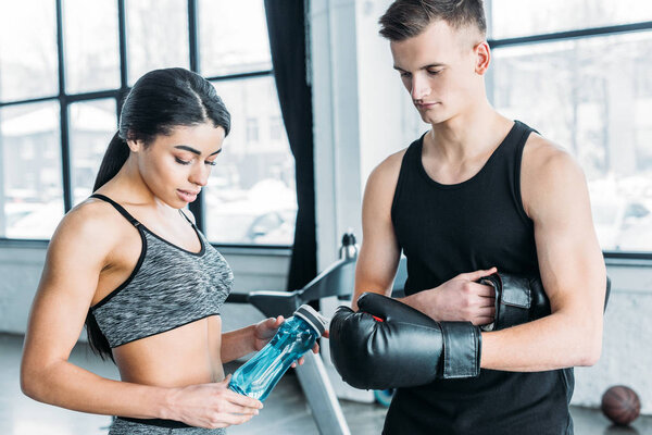 young man wearing boxing gloves and african american woman holding sports bottle in gym