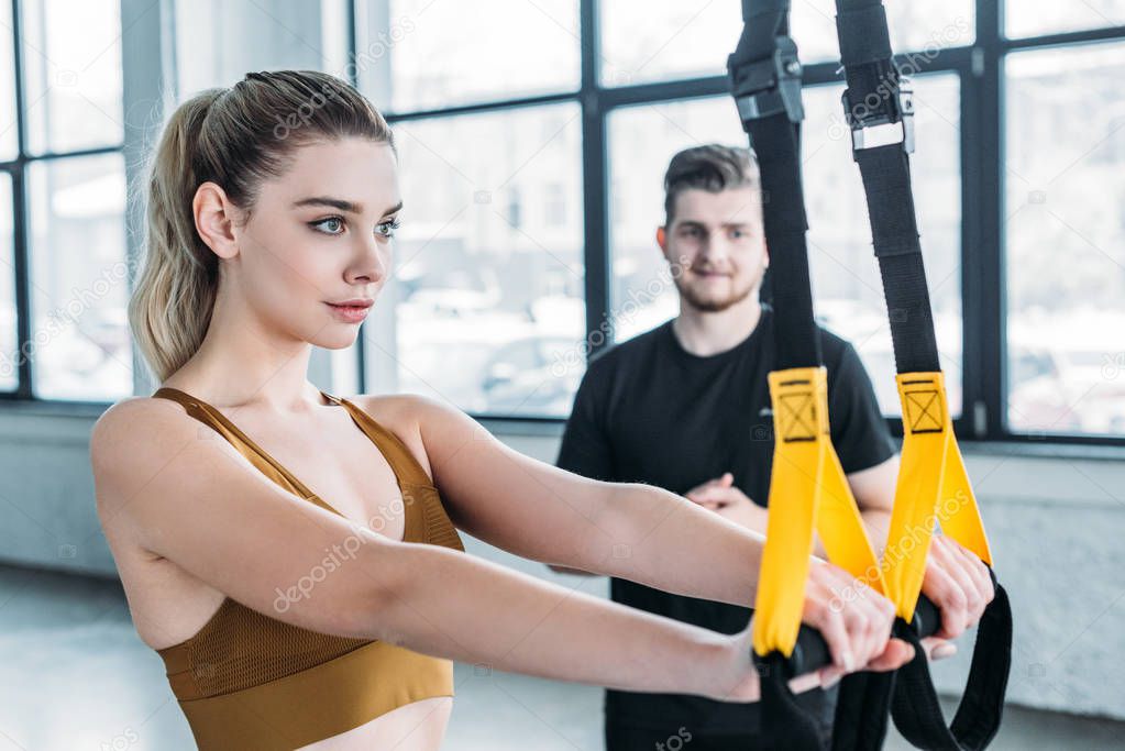 young man looking at beautiful sporty girl training with suspension straps in gym