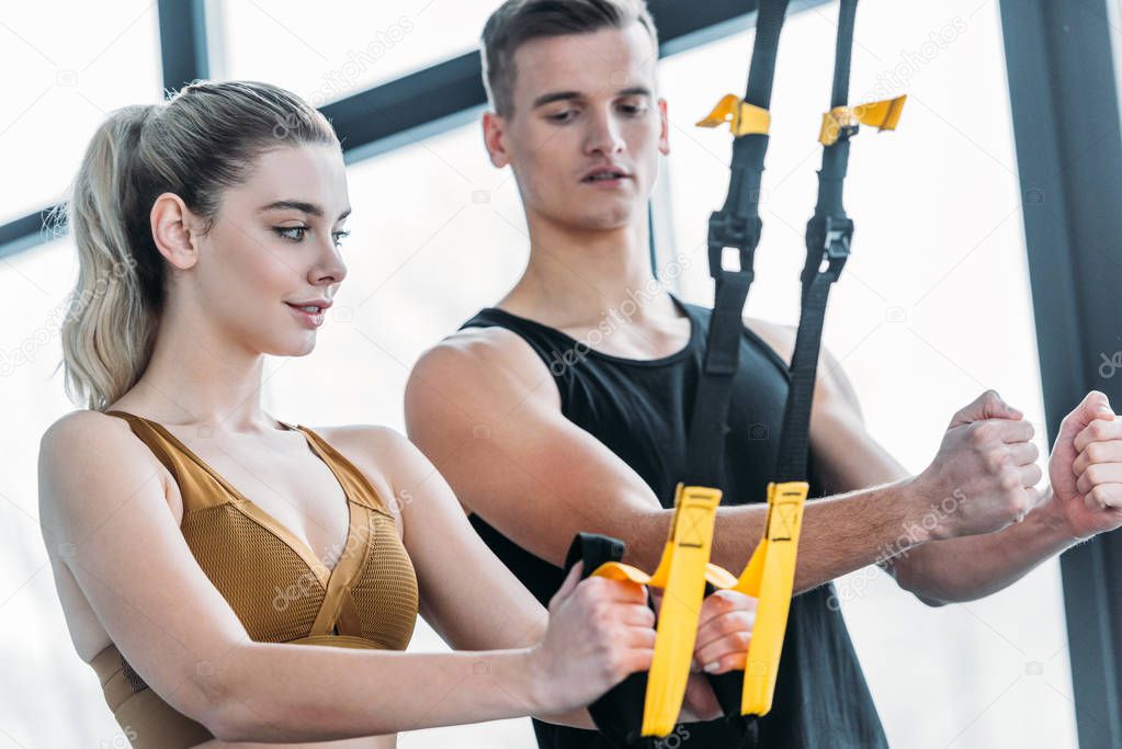 male trainer looking at beautiful sporty woman exercising with suspension straps in gym 