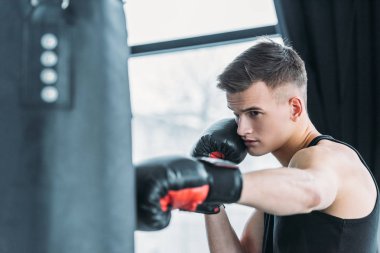 Spor salonunda kum torbasıyla boks yapan konsantre genç bir adam.