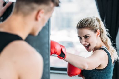 selective focus of male trainer looking at aggressive sportswoman boxing with punching bag in gym