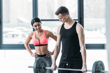 smiling african american girl in sportswear looking at handsome young sportsman lifting barbell in gym