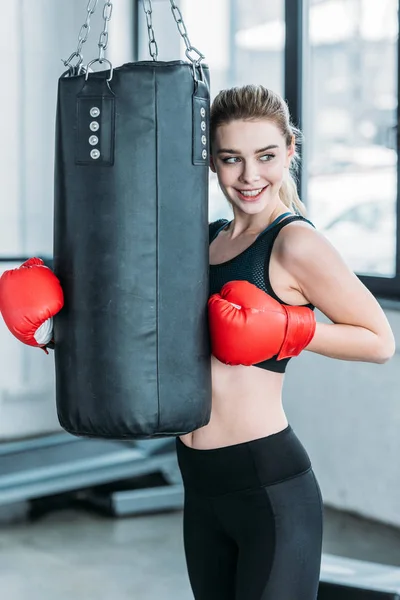 beautiful smiling young sportswoman in boxing gloves exercising with punching bag in gym