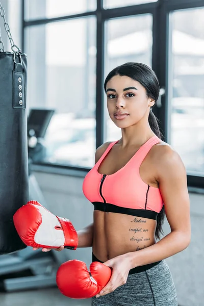 young african american sportswoman holding boxing gloves and looking at camera in gym