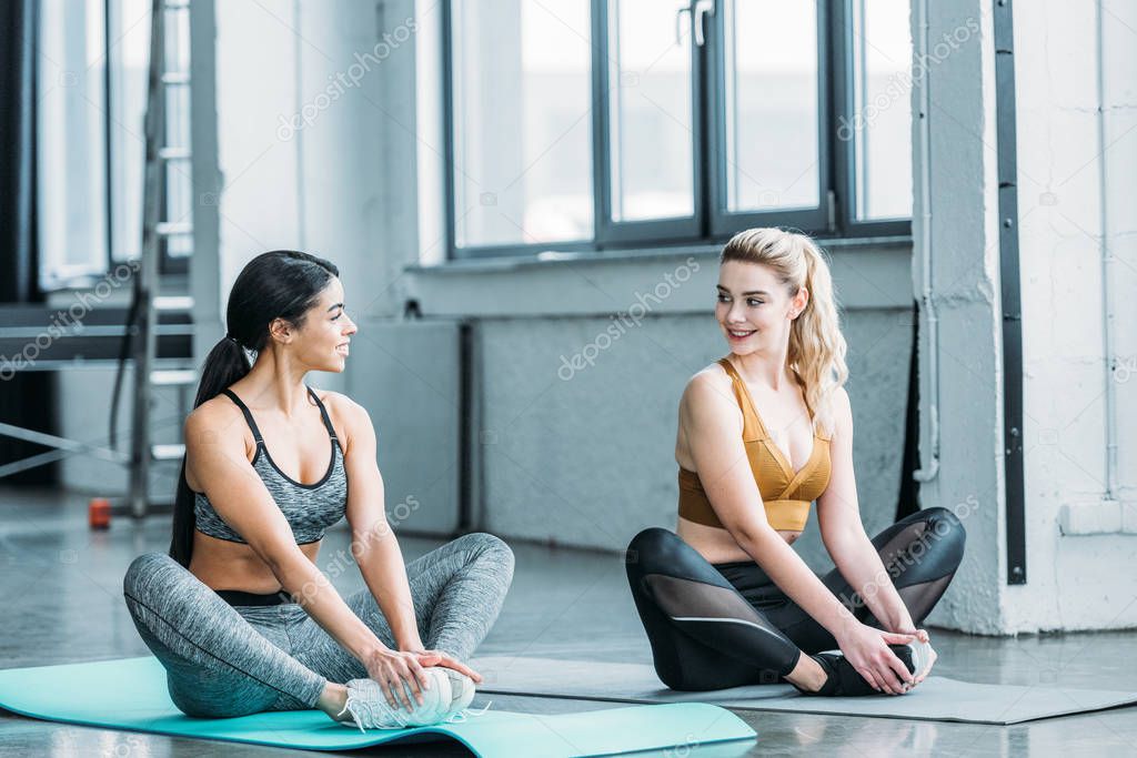 beautiful sporty girls sitting on yoga mats and smiling each other in gym