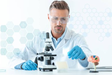 selective focus of handsome scientist in protective glasses making experiment with microscope isolated on white with medical symbols