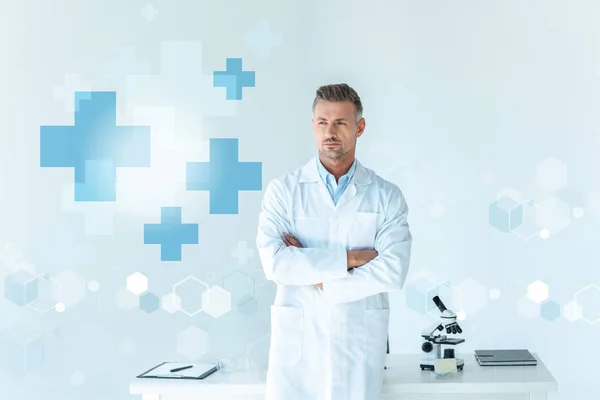英俊的科学家站在交叉的武器附近的桌子隔离在白色与医疗符号 — 图库照片