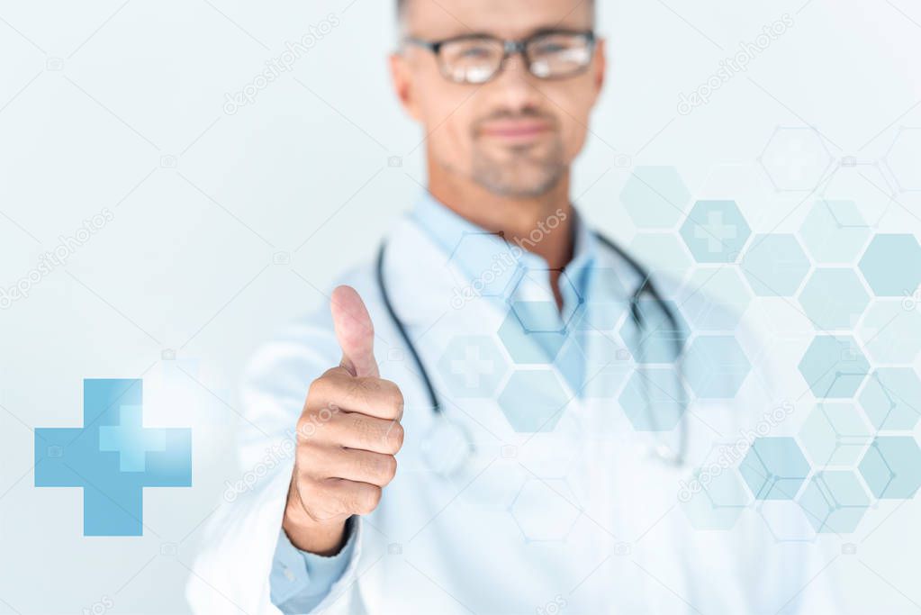 selective focus of handsome doctor in glasses with stethoscope on shoulders showing thumb up with medical interface