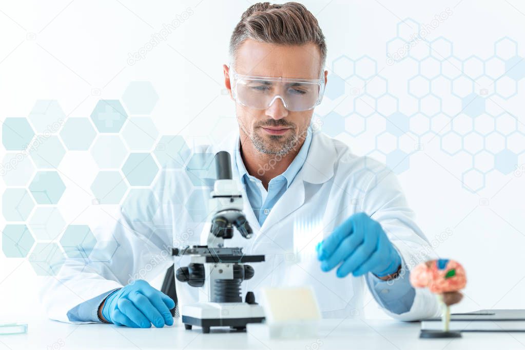 selective focus of handsome scientist in protective glasses making experiment with microscope isolated on white with medical symbols
