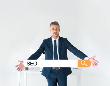 yakışıklı işadamı Grey seo arama çubuğunda ile izole bilgisayar ile tablo yakınındaki kollarını açarak ayakta