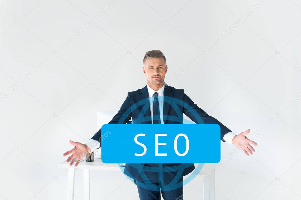 handsome businessman standing with open arms near table with computer isolated on grey with seo sign