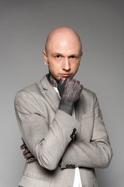 confident tattooed man in suit jacket standing with hand on chin and looking at camera isolated on grey clipart