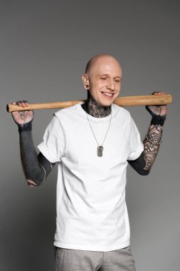 cheerful tattooed man standing with baseball bat on shoulders and laughing on grey  clipart