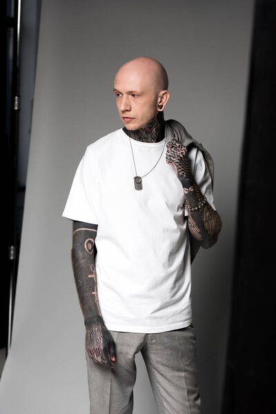 bald tattooed man in white t-shirt holding suit jacket on shoulder and looking away in studio