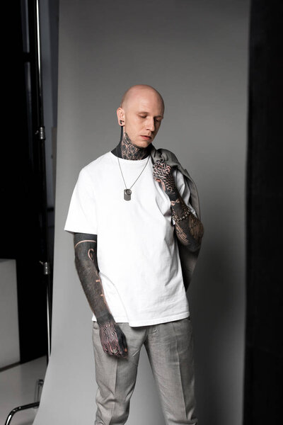 young bald man with tattoos holding suit jacket on shoulder and standing in studio
