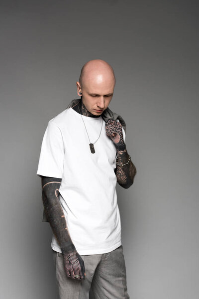 young tattooed man in white t-shirt holding suit jacket on shoulder and looking down isolated on grey