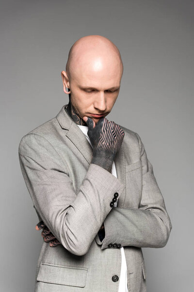 young tattooed man in suit standing with hand on chin and looking down isolated on grey